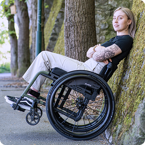 A young woman leans against a stone wall in her wheelchair.