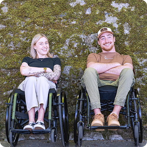 A young woman and a young man sit in wheelchairs in front of a moss-covered stone wall.