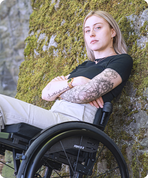 A young woman sits in her wheelchair against a stone wall.